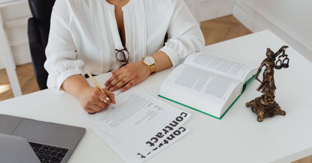 A legal professional in a black blazer signs a court summons document at a wooden desk with a pen.