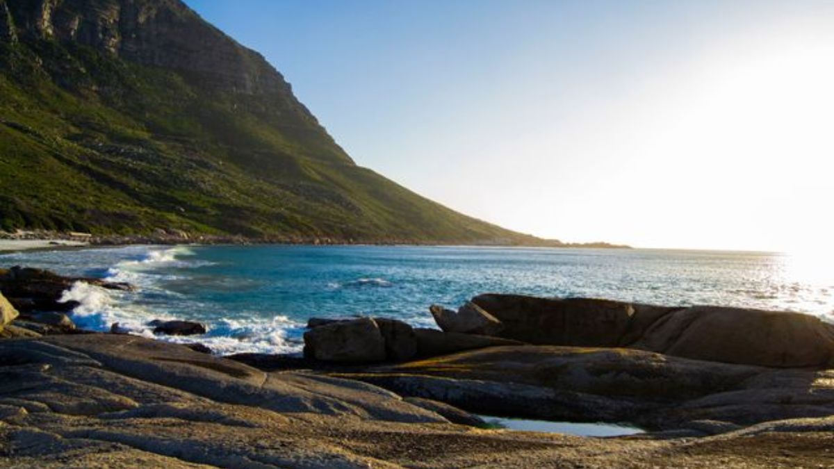 Wooden boardwalk leading to white sand beach and turquoise water at Llandudno Beach in Cape Town with coastal mountain background.
