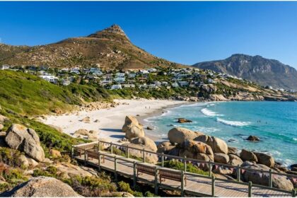 Wooden boardwalk leading to white sand beach and turquoise water at Llandudno Beach in Cape Town with coastal mountain background.