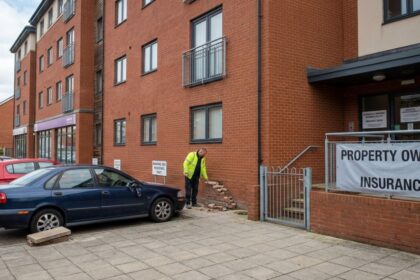 Damaged red brick building exterior next to a property owners insurance banner and parked cars.