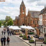 Pedestrians browsing an outdoor street market in Rugby England town surrounded by historic brick buildings.