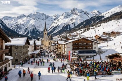 Snowy Alpine ski village of St. Anton featuring traditional chalets, a church steeple, ski lifts, and people gathered outdoors under majestic, snow-capped mountain peaks.