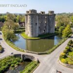 Nunney Somerset England, featuring a medieval stone castle surrounded by a moat, village houses, trees, and countryside.